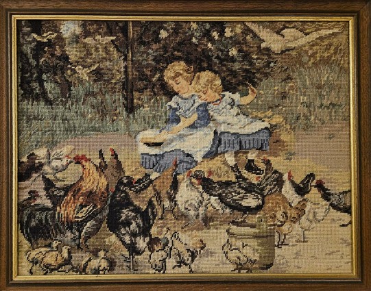 A crossstitch picture in a frame showing two small girls feeding chickens.